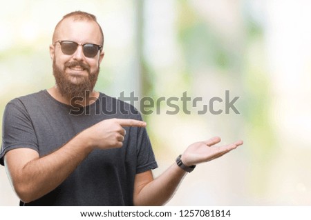 Young caucasian hipster man wearing sunglasses over isolated background amazed and smiling to the camera while presenting with hand and pointing with finger.
