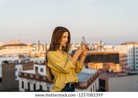 pretty young hipster woman in a yellow shirt taking picture with cell phone. charming beautiful girl making photo with gadget. happy lady on a rooftop during sunset 