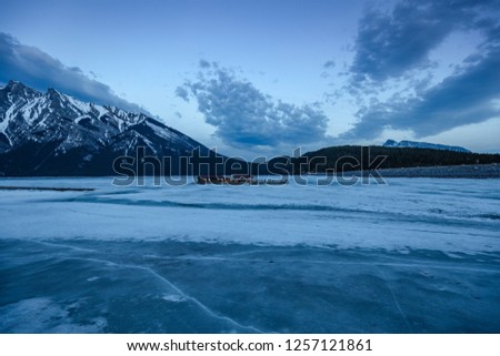 Lake Minnewanka located in Banff, Canada. Glacial lake frozen during winter. Blue hour at the lake with glacial mountain and cloud background. House in the middle of a frozen lake.