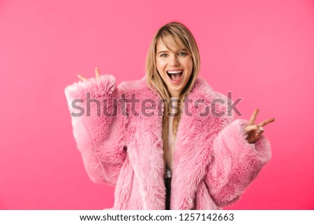 Cheerful young blonde woman wearing sweater standing isolated over pink background, wearing pink fur coat
