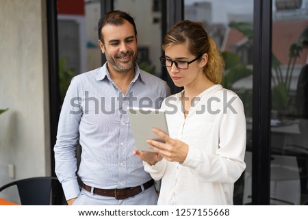 Serious female trainee showing her work to executive on digital tablet. Mid adult Caucasian businessman looking at pc and smiling. Apprentice concept