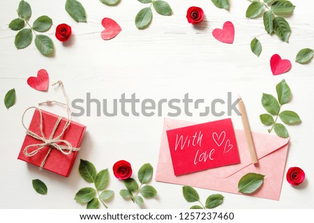 Red card with an inscription "With love", hearts, gift box and pink envelope on a white wood background, top view. Romantic background by St. Valentine's Day