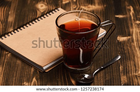 Tea and notebook on wooden background. Composition of cup and textbook for relax. Autumn evening concept. Glass of black tea with tea bag near tea spoon and closed book.