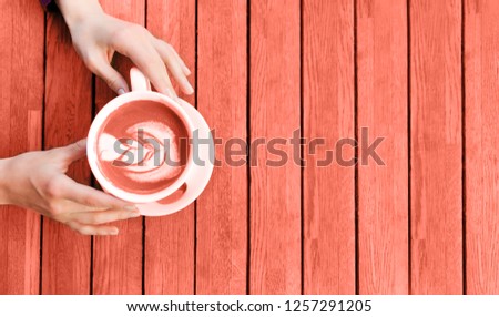 Woman's hands holding cup of cappuccino on wooden table. Coral colored picture. Beverage concept of color of the year 2019. Top view, flat lay, copyspace for text