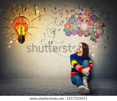 Young woman sitting on the floor smiling looking at a bright light bulb on the wall. Idea concept, creative thinking as colorful gear brain above head, create a genius. Mental development symbol. Royalty-Free Stock Photo #1257319651
