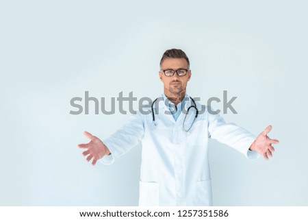handsome doctor in glasses with stethoscope on shoulders standing with open arms and looking at camera isolated on white