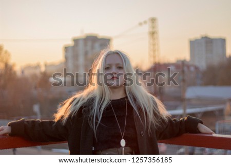 girl with blond hair and green eyes stands on a bridge in a coat at sunset