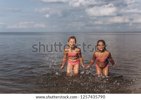 Drops of water are doing by two happy cheerful caucasian girls having fun on the coast of ocean in summer. Children are on background, blurred. Happiness, summer vacation, childhood concept