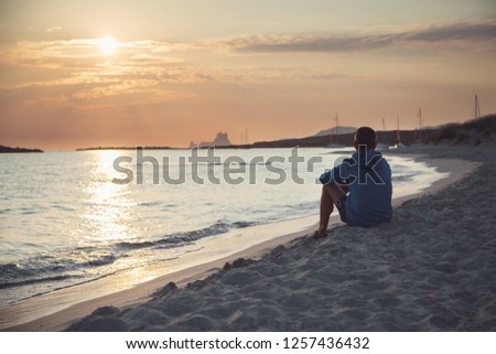 A young man sits on the beach in the sand and looks into the distance.