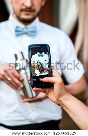 Girl takes photo a bartender with a shaker. Focus on the smartphone. Bearded barman in a bow tie makes a cocktail. Royalty-Free Stock Photo #1257461329