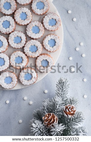 Flower Linzer cookies with blue icing on white marmor board set on top of light winter background with Christmas tree decorated with pine cones