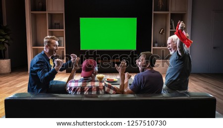 A group of fans is watching a TV and celebrating some joyful sports moment, sitting on the couch in the living room. The living room is made in 3D. TV is green screen for further editing.