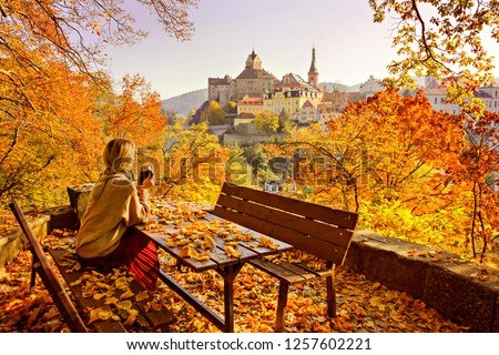 Blond girl with mug of hot steamy beverage sitting on a bench strewn with fallen yellow leaves, enjoying at the colorful landscape. Concept of enjoying  nature, life, relaxation, travel.  Royalty-Free Stock Photo #1257602221