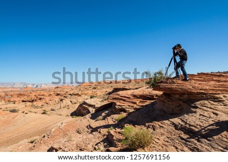 Young man stands and takes a photo with a tripod at the edge of red rock, Grand Canyon National Park, Arizona-USA. A man taking pictures with blue sky at Horseshoe Bend, Arizona-USA.