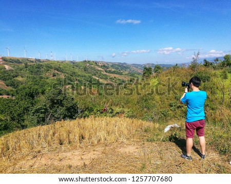 Back view of one photographer that he is taking a picture of high scenery valley in Thailand.  There are white wind turbine on top of mountain at far view. It is a sunny day with clear blue sky.