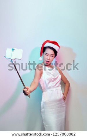 Modern holiday concept. Blonde woman in Santa hat taking Christmas picture of herself, selfie with smartphone on stick.