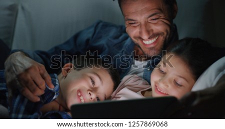 Portrait of happy father and kids using a tablet on sofa in the evening. Concept of family entertainment, education, technology