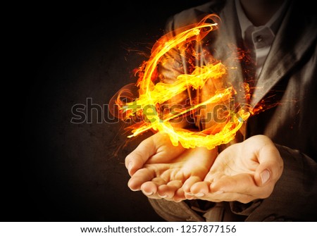 Close-up of businessman in suit presenting flaming euro sign in his hands with dark wall on background.