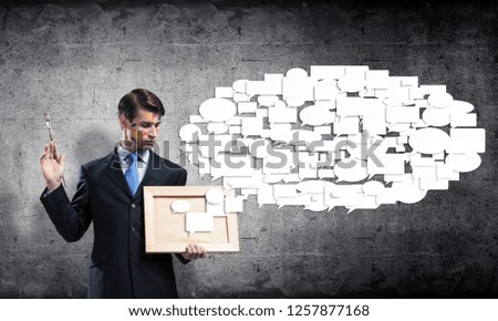 Young and successful businessman in black suit holding paintbrush in hands and looking at picture frame while standing against gray concrete wall on background