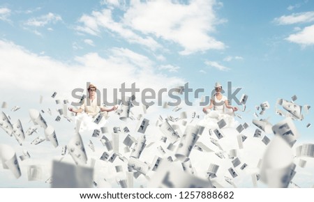 Young couple keeping eyes closed and looking concentrated while meditating among flying papers in the air with cloudy skyscape on background.