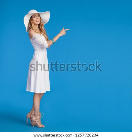 Beautiful young woman in white summer dress, high heels and sun hat is standing, pointing and looking at camera, Side view. Full length studio shot on blue background.