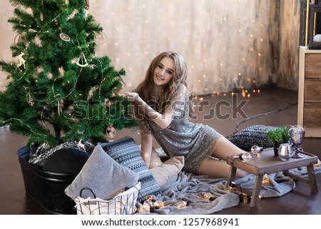 Beautiful girl sitting near the Christmas tree in a silver dress with sequins. Sits on the floor on a plaid with pillows. Holds a Christmas tree branch. New Year's interior.
