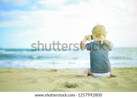 Little girl is taking photo by phone at the beach seating on the sand. back view photo.  