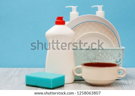 Plastic bottles of dishwashing liquid, glass and tile cleaner, clean plates in basket, bowl and sponge on blue background. Washing and cleaning concept.