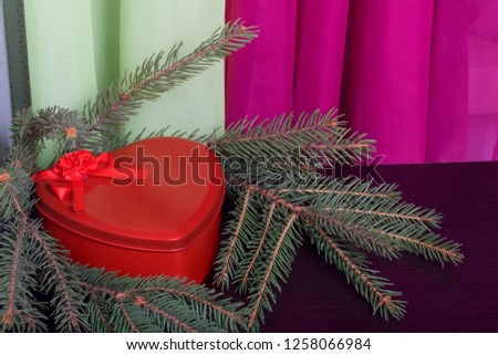 Red box with a gift. Made in the shape of a heart. Near the fir branches. Against the backdrop of curtains of lime and lilac color.