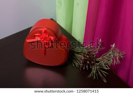 Red box with a gift. Made in the shape of a heart. Near pine branch with a cone. Against the backdrop of curtains of lime and lilac color.