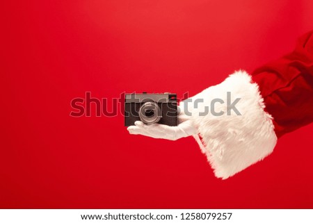 The hand of santa claus holding a camera on red background. The season, winter, holiday, celebration, gift concept