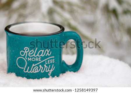 Blue hot mug steam rising relax more worry less on front surrounded by snowy scene & icy pine branches winter background, holiday stress keep calm relax more worry less concept, text copy space