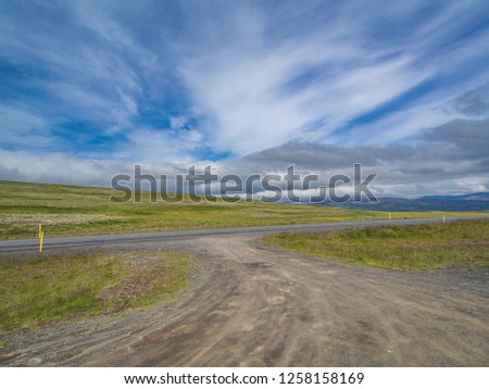 Crossroads between asphalt road and dirt road. Adjoining minor road to main road. Beautiful icelandic rural landscape with green meadows under blue sky with clouds. Summer day, panoramic view.