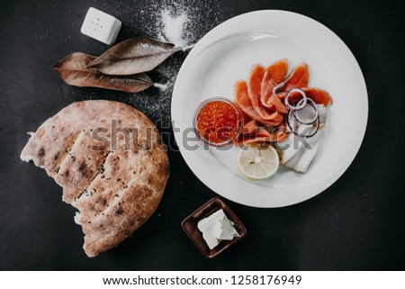 Salmon slices with butterfish slices, lemon and a red caviar on the white plate with a butter, bread, salt and magnolia leaves nearby.