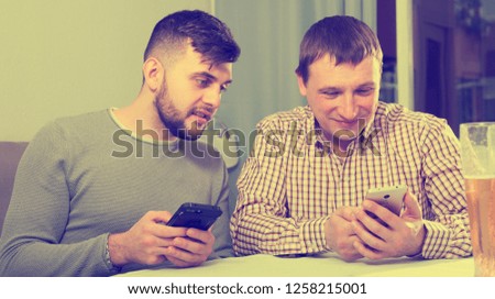 Happy male friends using phone conversing with liveliness at home table