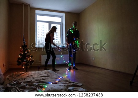 Girl wraps guy in multi-colored garland in a large room.