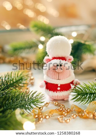 Handmade knitted toy. Christmas pig in Santa Claus costume in light christmas background with red berries, branches  and lights