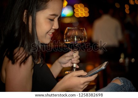 Red Wine Party.  Happy Young Woman Having a Glass of Red Wine while Using Smartphone Checking Social Online Against Blurred Bokeh Nightclub. Picture Presenting Happy Beautiful Girl with Red Wine
