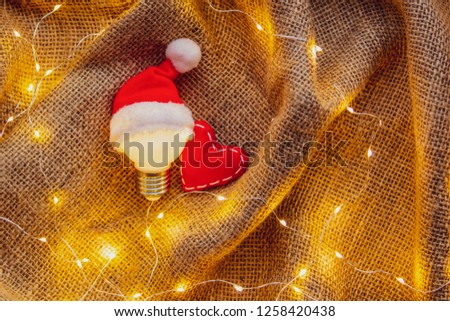 bulb in Christmas hat and heart shape toy on jute background