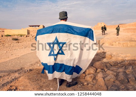 Israeli military infantry stands in middle of the desert holding an Israeli flag with the star of David. Jewish patriot. Military exercises at Masada Fortress on mountain in Judean desert in Israel. Royalty-Free Stock Photo #1258444048
