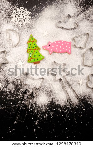 Christmas composition with gingerbread in the shape of a new year tree and a pig, baking molds for cookies, white plastic snowflakes and artificial snow on a black background. Copy space down below.