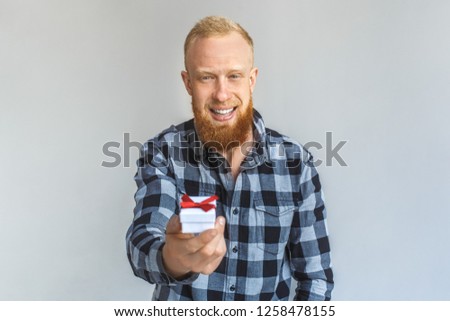 Red hair mature man standing isolated on grey wall giving small gift box looking camera smiling happy