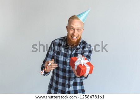 Red hair mature man wearing birthday cap standing isolated on grey wall holding gift box and party horn looking camera laughing playful