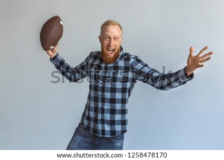 Red hair mature man standing isolated on grey wall throwing american football ball looking camera shouting excited