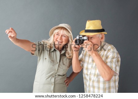Senior couple tourists wearing beach hats studio standing isolated on gray wall holding camera taking photos choosing composition smiling cheerful