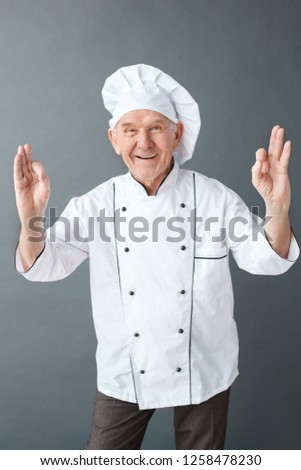 Senior male chef studio standing isolated on gray wall looking camera smiling cheerful showing ok sign with both hands