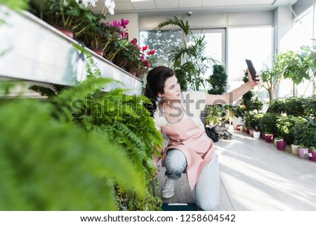 Young modern beautiful female florist taking selfie with her smartphone for social media in botanical garden