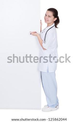 Smiling young woman doctor pointing to a large blank white sign or placard with copyspace for your text or advertisement