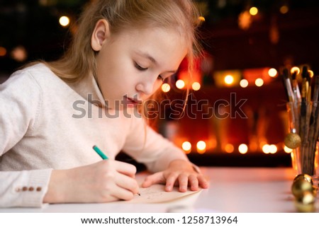 Blonde child girl writing letter to the Santa Claus or drawing something on the background with warm yellow bokeh. Christmas and New Year theme.