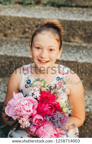 Summer portrait of adorable little girl wearing beautiful occasional dress, holding big peonies bouquet, posing outside